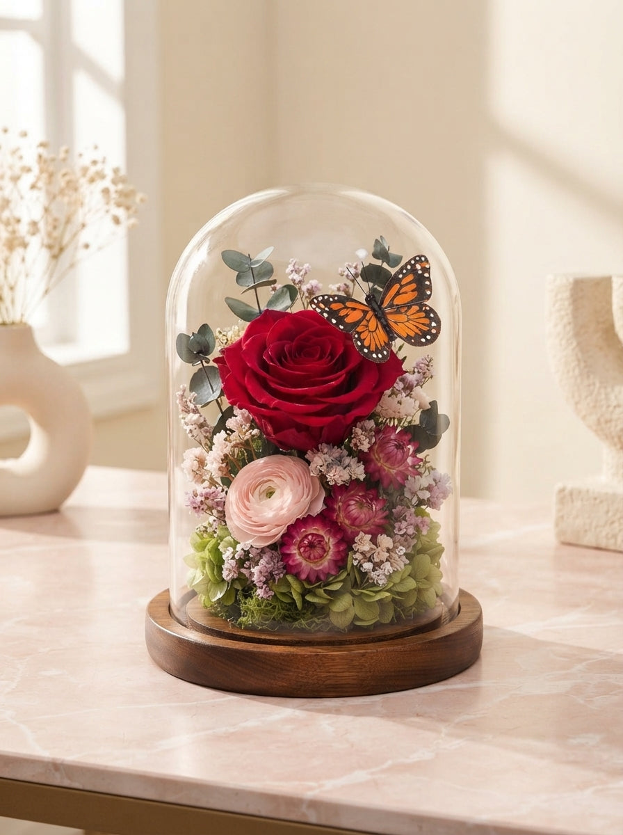 A preserved red rose in a glass dome on a wooden base sitting on a marble surface in a sunlit room.