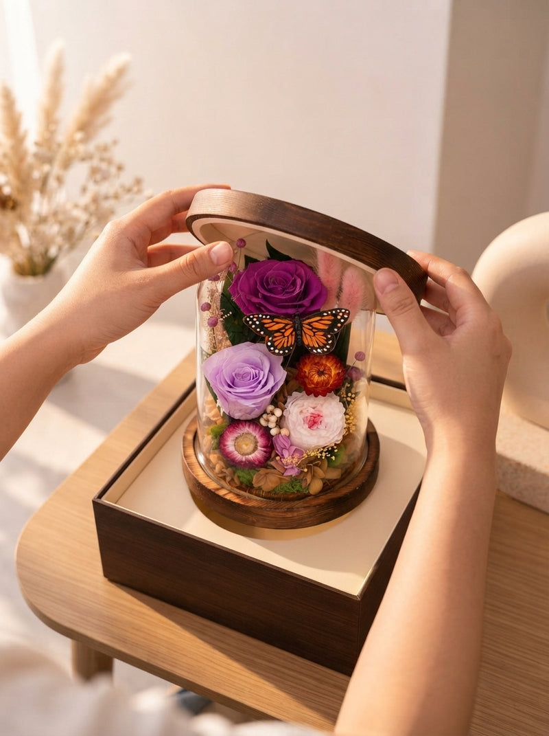 Hands opening a gift box to reveal a preserved rose inside a glass dome on a wooden table.