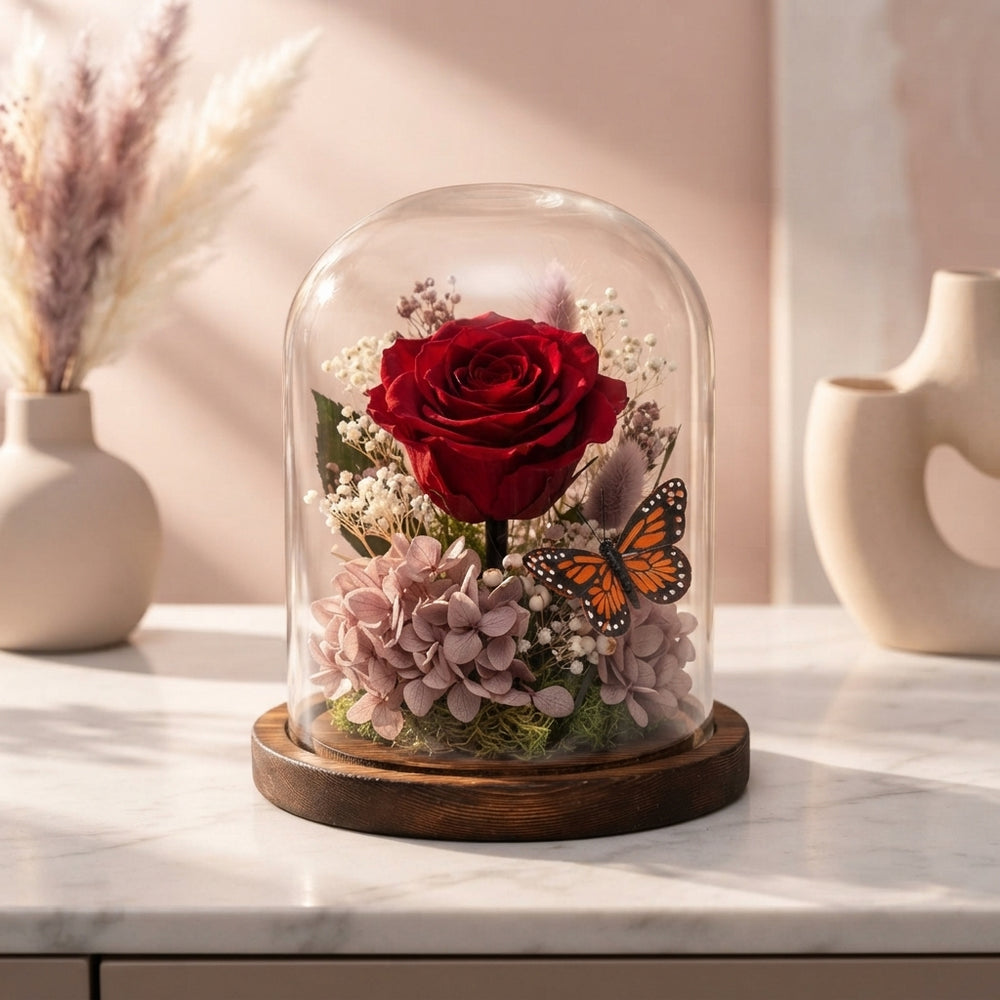 A preserved red rose inside a glass dome displayed on a bedside table in a modern, elegant bedroom with soft pink and cream t
